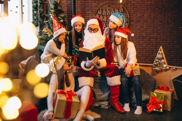 聖誕老人坐在沙發上，周圍圍繞著五個戴著聖誕帽的孩子，他們一起欣賞著一本書，背景有裝飾的聖誕樹和節日裝飾，還有許多禮物袋與禮物。